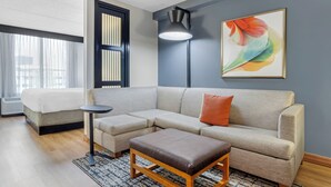 Une chambre d’hôtel moderne, dotée d’un canapé d’angle, d’une petite table d’appoint, d’un lit simple et d’une grande fenêtre avec des stores.