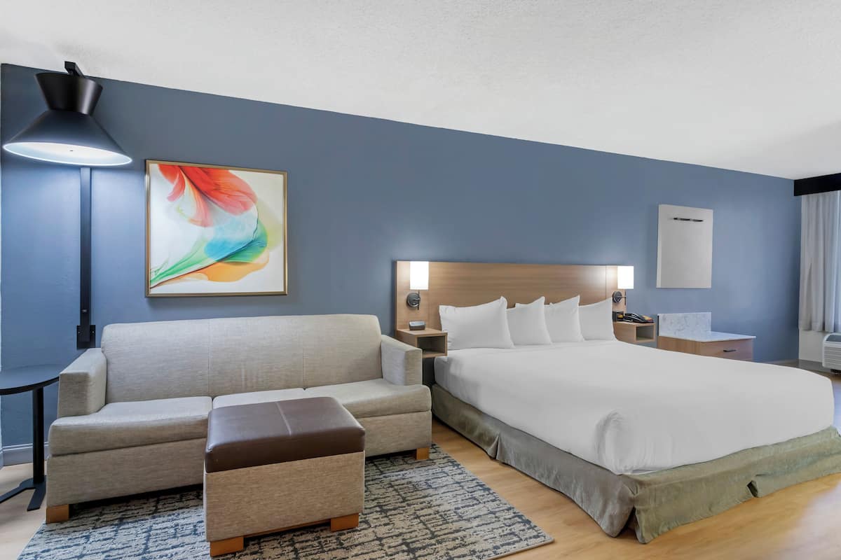 A hotel room with a bed, a sofa, a nightstand, a lamp, and a painting.
