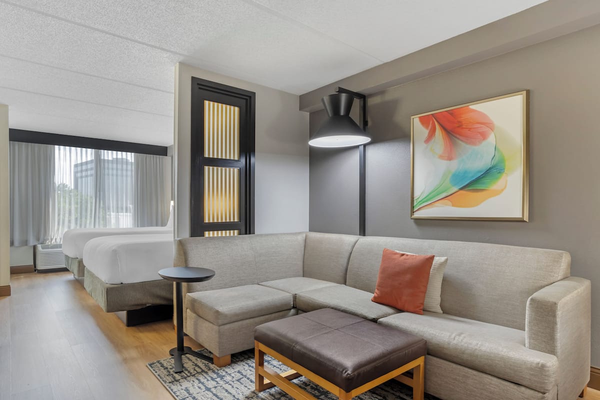 A modern hotel room with a sectional sofa, a small table, a bed with curtains, a floor lamp, and a colorful abstract painting.