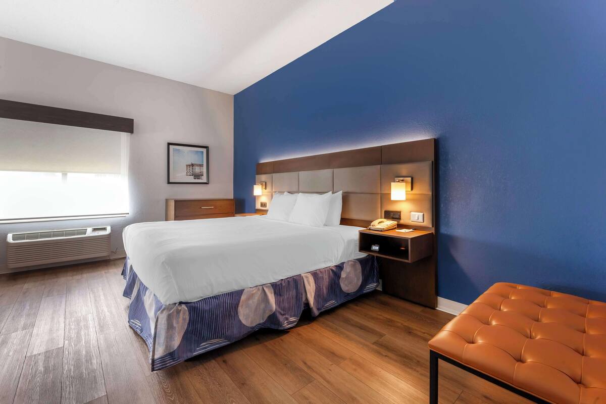 A hotel room with a large bed, a bedside table with a lamp, a wooden bench, and a window with blinds.