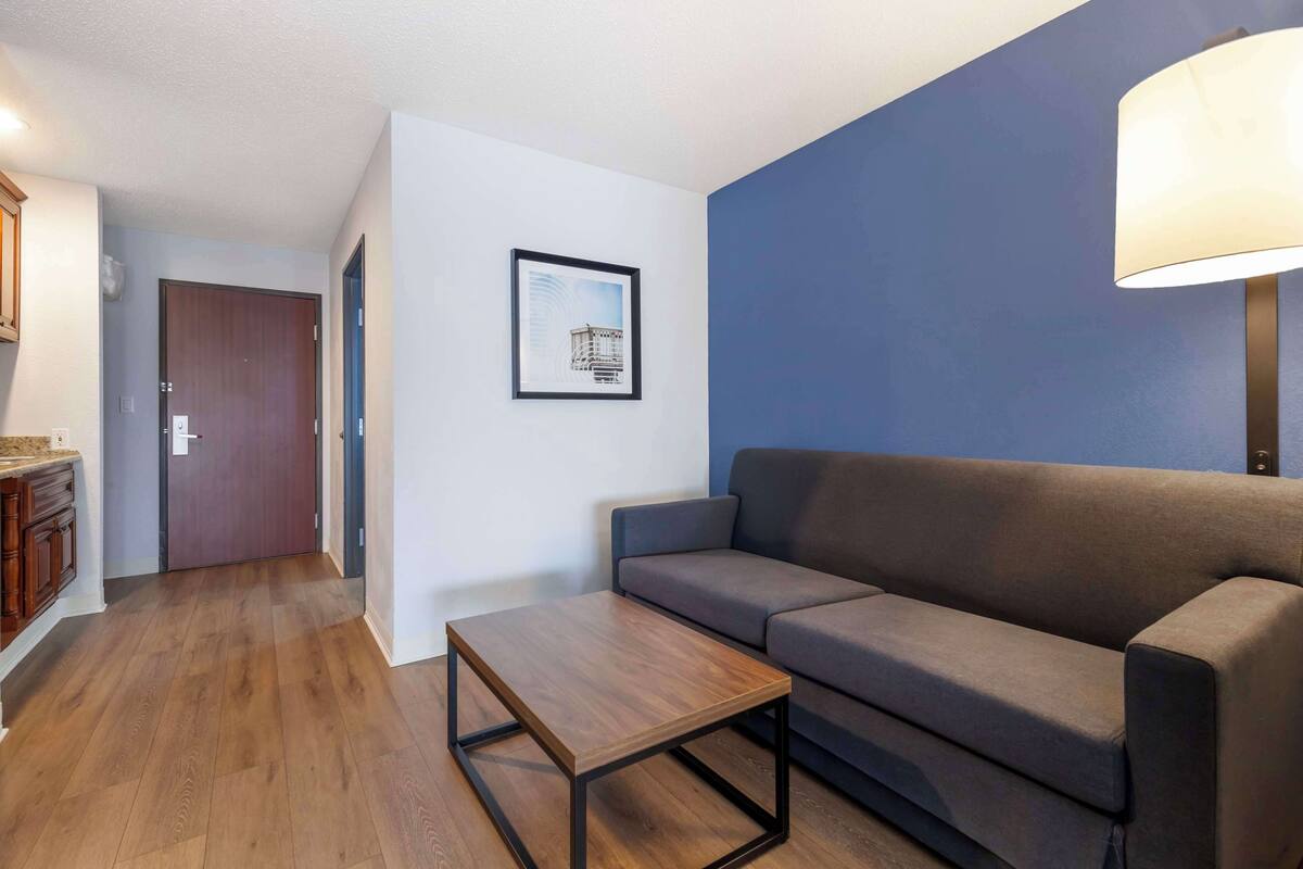 A modern living room with a wooden floor, a grey sofa, a small wooden table, a framed picture on the wall, and a lamp on the right.