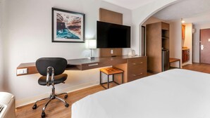 A hotel room with a bed, a desk with a chair, a television, a small stool, and a framed picture on the wall.