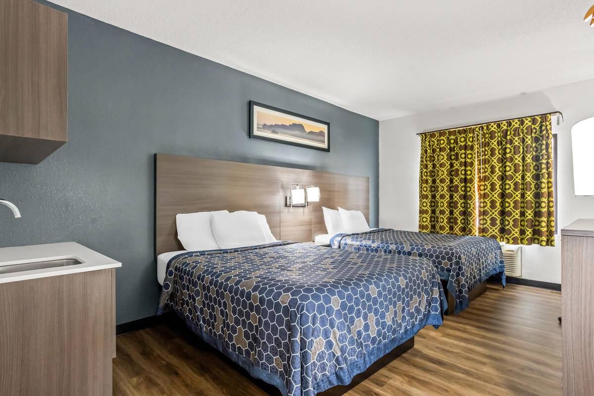 Ein Hotelzimmer mit zwei Betten, einem Holzkopfende, einem Bild an der Wand und einem Fenster mit gemusterten Vorhängen.