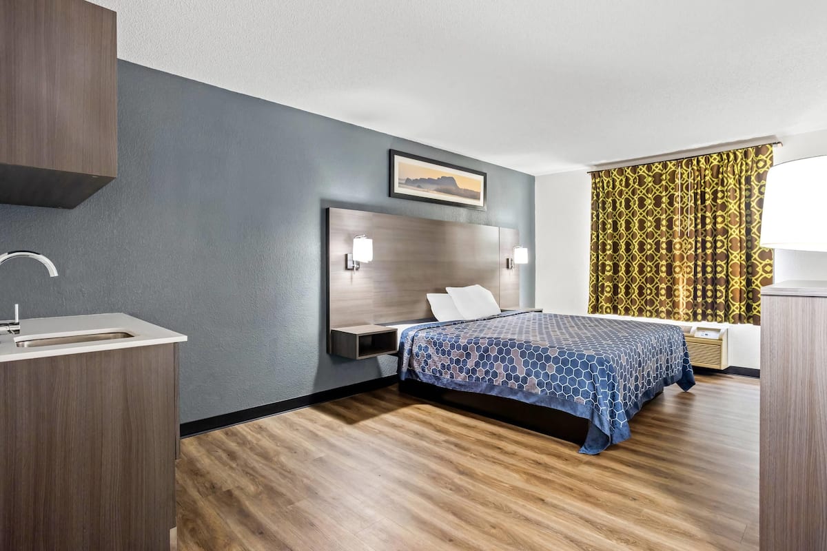Ein modernes Hotelzimmer mit einem großen Bett, einem Nachttisch, einem eingebauten Schreibtisch mit Waschbecken und einem gemusterten Vorhang.