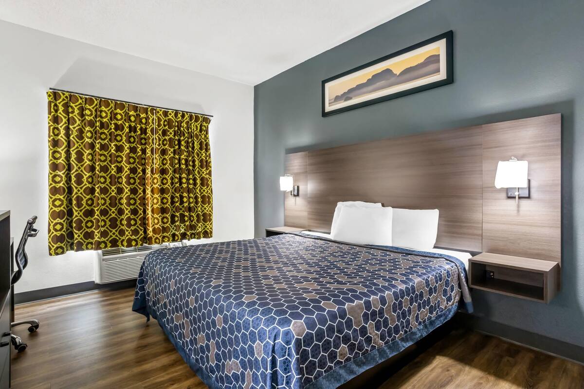 Ein Hotelzimmer mit einem großen Bett, einem Kopfteil mit integrierter Beleuchtung, einem Fenster mit gemusterten Vorhängen und einem Gemälde an der Wand.