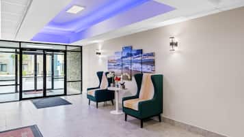 A lobby with a glass entrance, teal armchairs, a small round table with a vase of flowers, and a wall-mounted artwork.