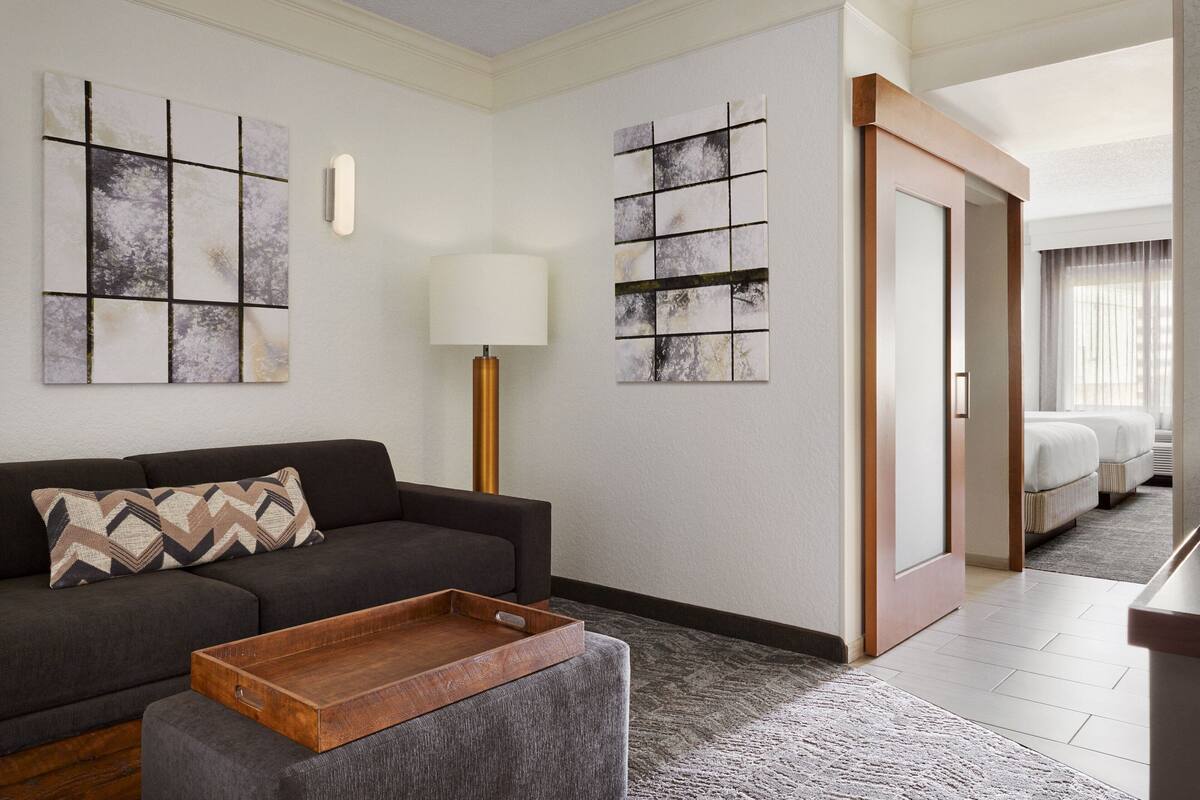 Une chambre d’hôtel moderne avec un canapé gris, une table d’appoint en bois et des œuvres d’art abstraites accrochées au mur.