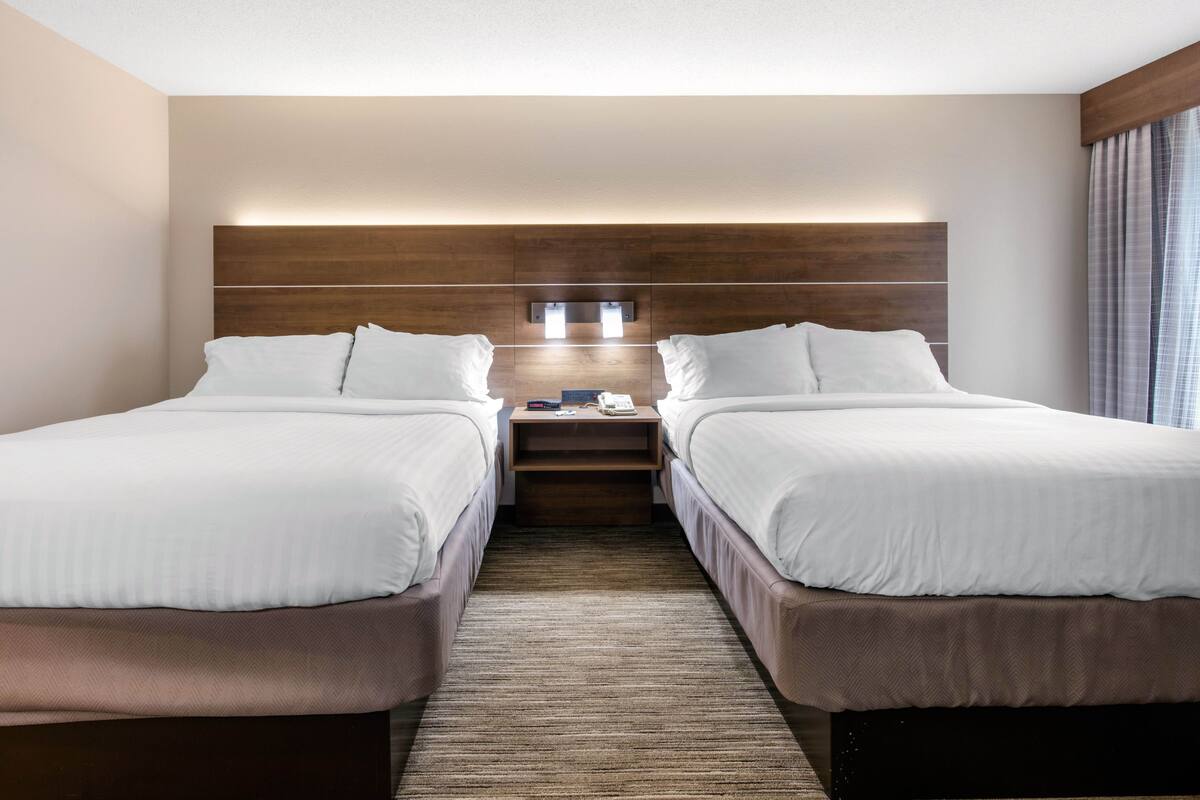 Habitación de hotel con dos camas, cabecera de madera, mesita de noche con teléfono y ventana con cortinas.