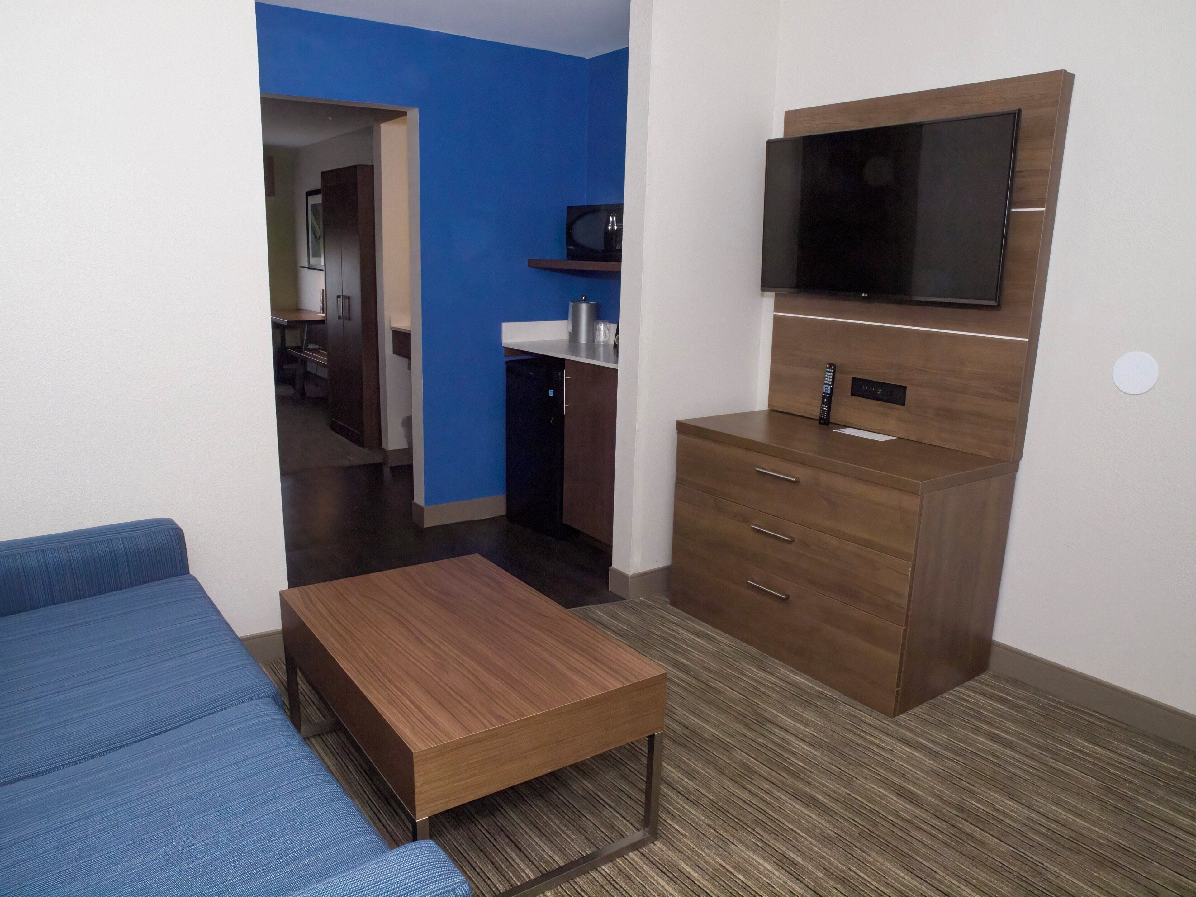 A compact room with a sofa, a small wooden table, a TV mounted on the wall, and a kitchenette area.