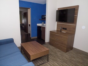 A compact room with a sofa, a small wooden table, a TV mounted on the wall, and a kitchenette area. - Holiday Inn Express Hotel & Suites Madison by IHG (Madison)