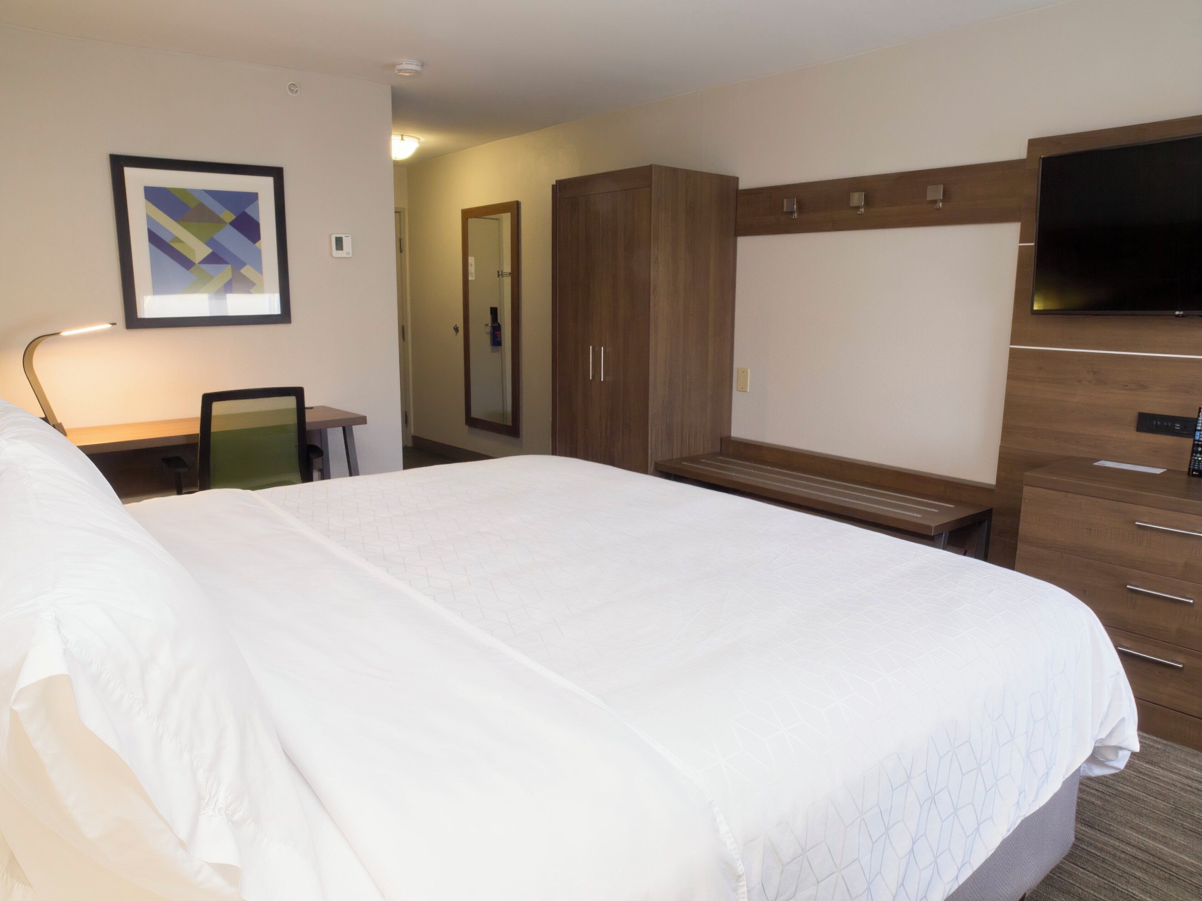 A hotel room with a large bed, a desk with a chair, a wardrobe, a television, and a framed picture on the wall.