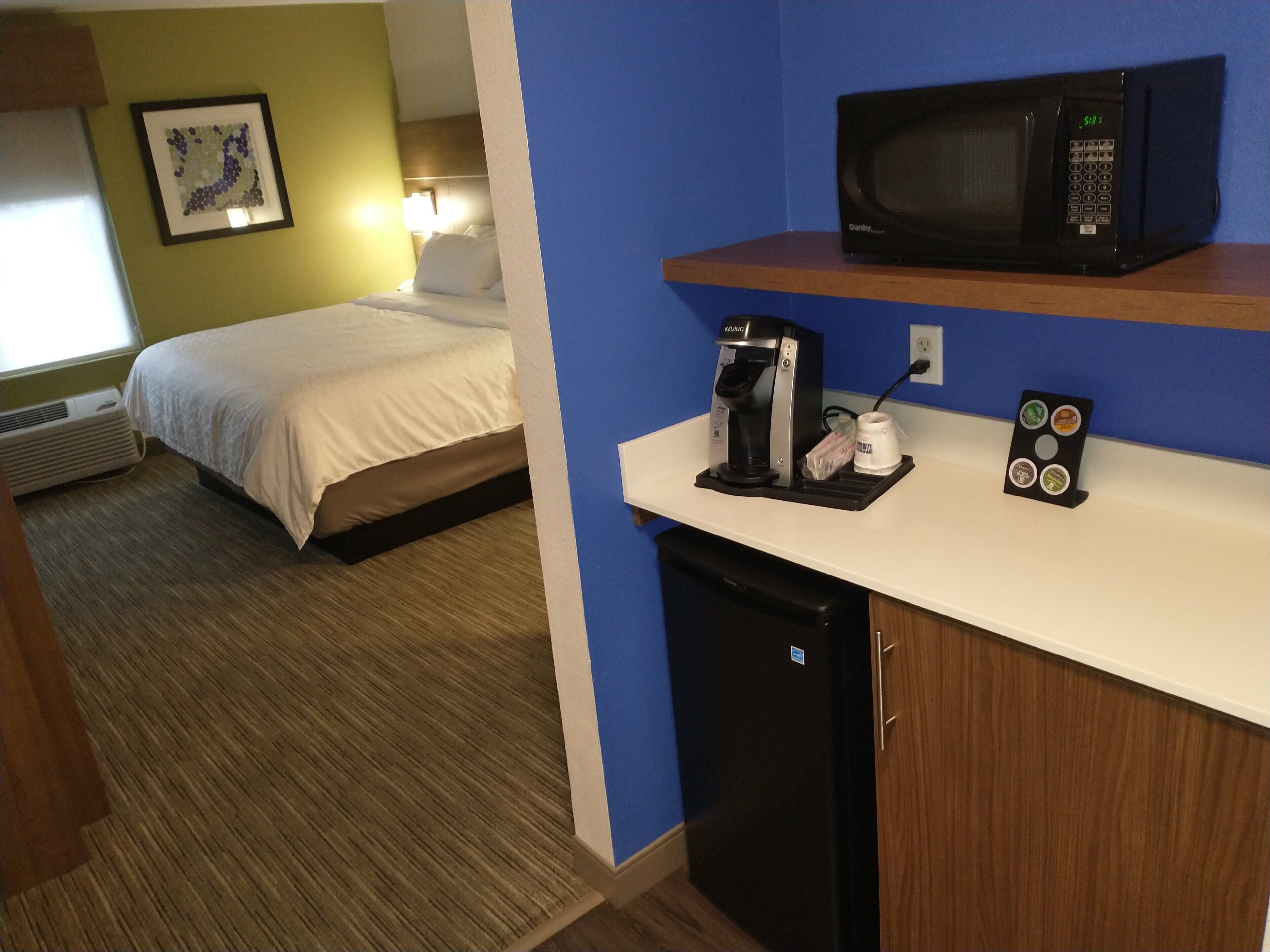 Habitación de hotel con cama, cafetera, microondas y refrigerador pequeño.