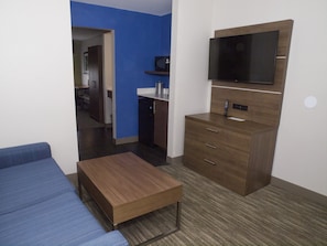 Habitación de hotel compacta con sofá, una mesita de madera, kitchenette y televisor montado en la pared.