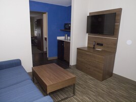 Habitación de hotel compacta con sofá, una mesita de madera, kitchenette y televisor montado en la pared.