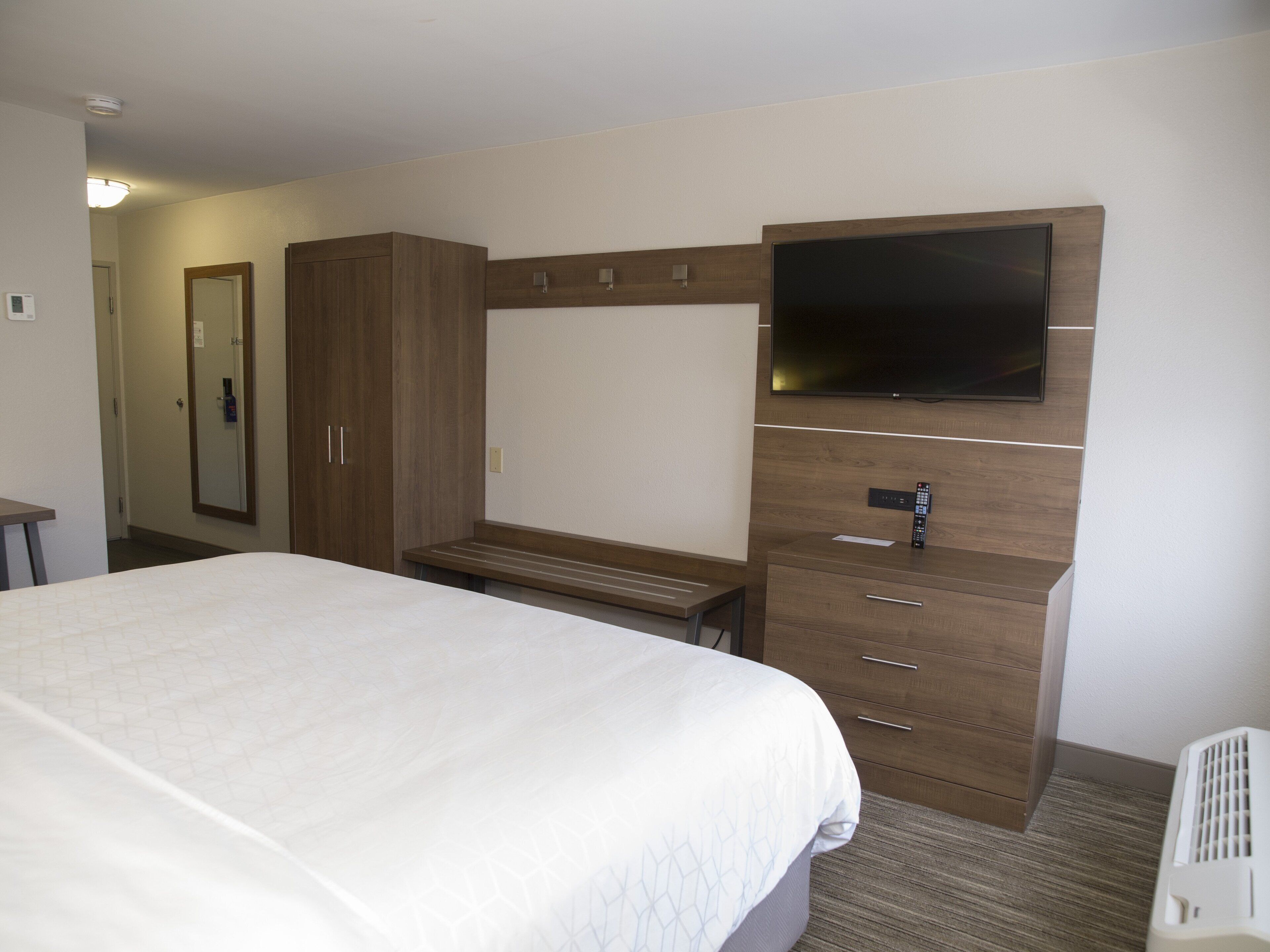 A hotel room with a bed, a TV mounted on the wall, a wooden wardrobe, and a desk.