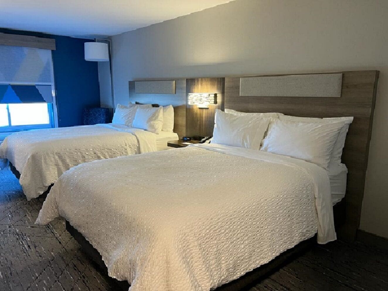 Habitación de hotel con dos camas, cabecero de madera, mesita de noche y ventana con persianas.