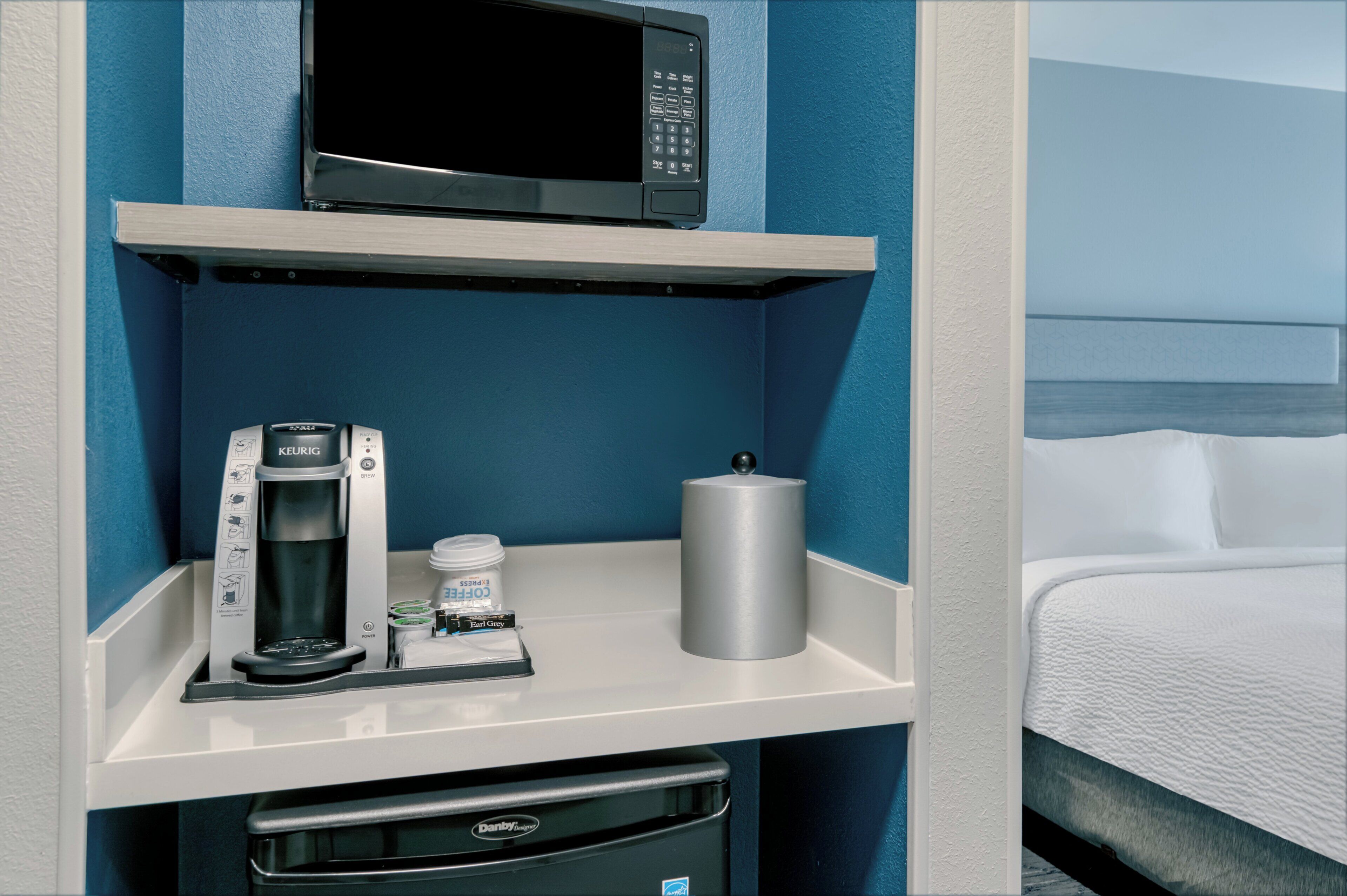 A hotel room with a microwave, coffee maker, and a container of coffee pods on a white shelf.