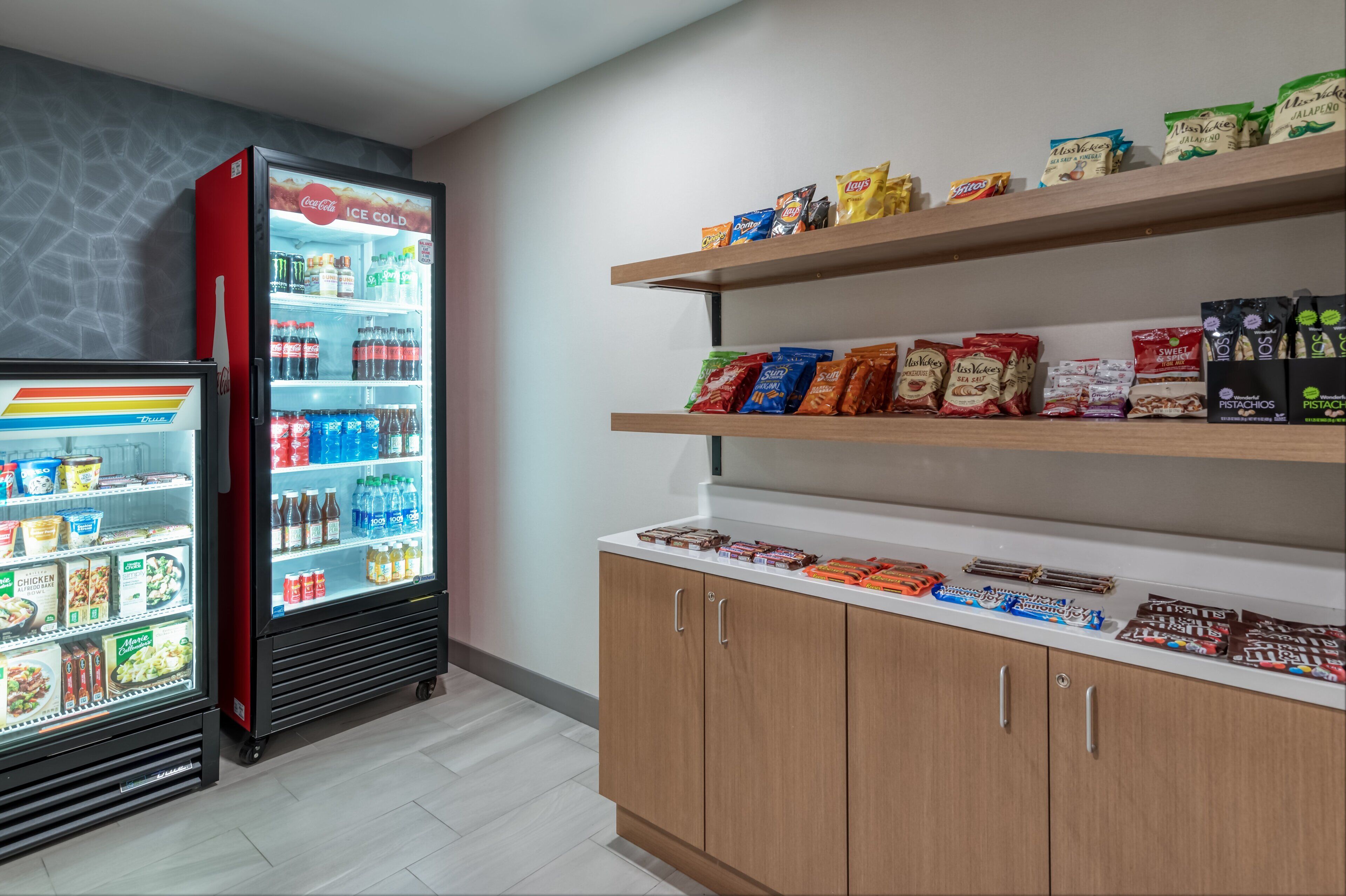 A refrigerated display with beverages and snacks, wooden shelves with packaged goods, and a white countertop with chocolates and other items.