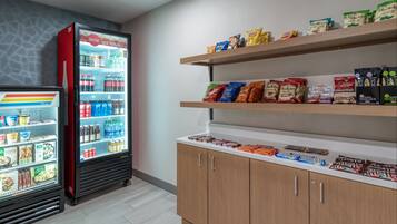 A refrigerated display with beverages and snacks, wooden shelves with packaged goods, and a white countertop with chocolates and other items.