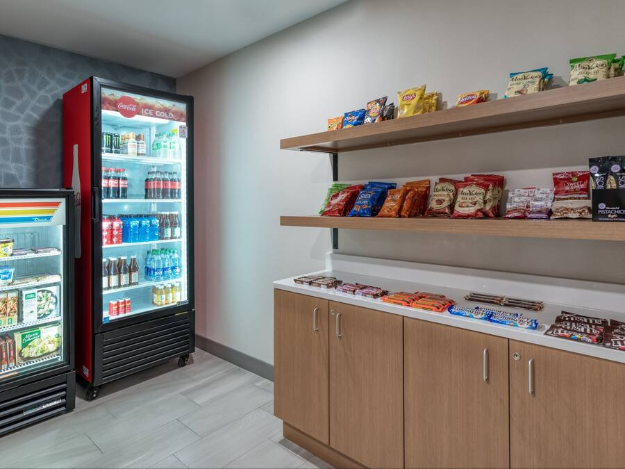 A refrigerated display with beverages and snacks, wooden shelves with packaged goods, and a white countertop with chocolates and other items.