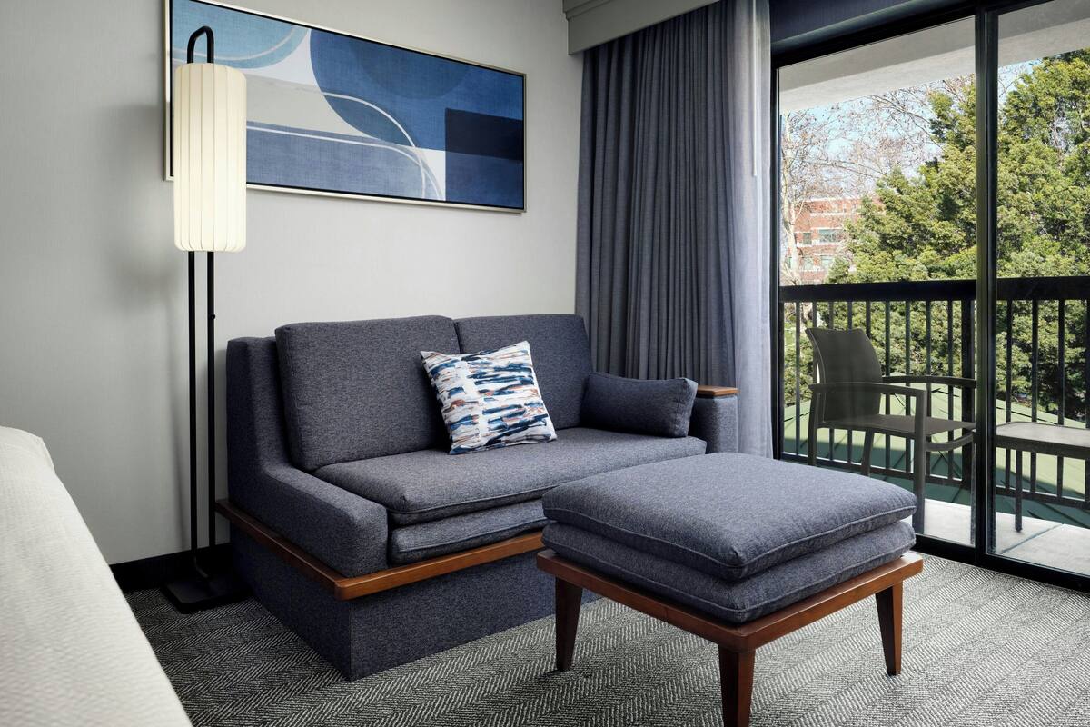A modern hotel room with a grey sofa, a matching ottoman, a floor lamp, and a large window offering a view of trees and buildings.