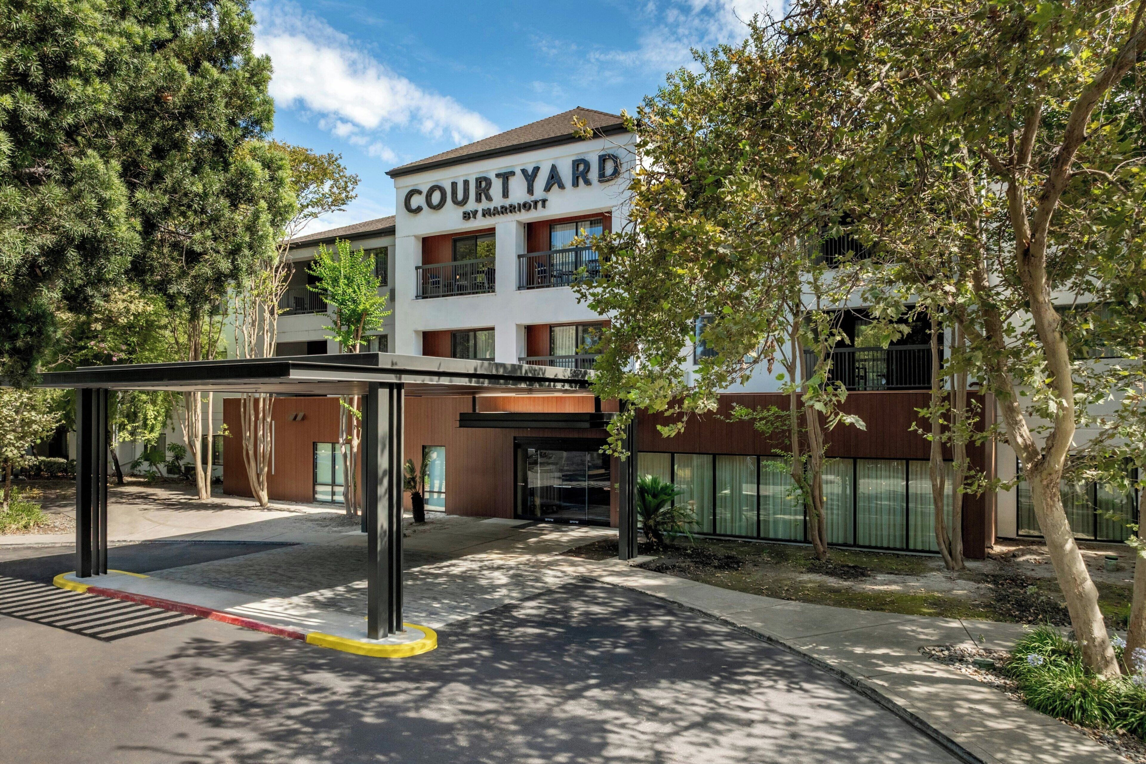 A Courtyard by Marriott hotel entrance with a covered walkway, trees, and a clear sky.