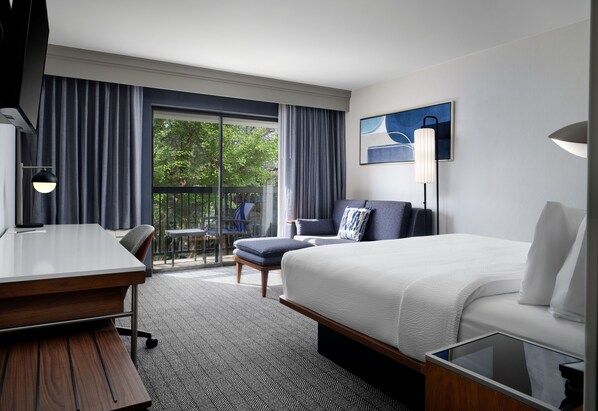 A hotel room with a bed, a desk, a chair, a sofa, a large window, and a painting on the wall.