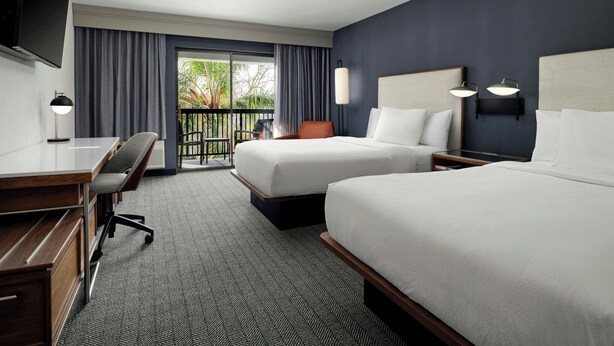 A hotel room with two beds, a desk, a chair, a TV, and a balcony with curtains. - Courtyard by Marriott Stockton (Stockton)