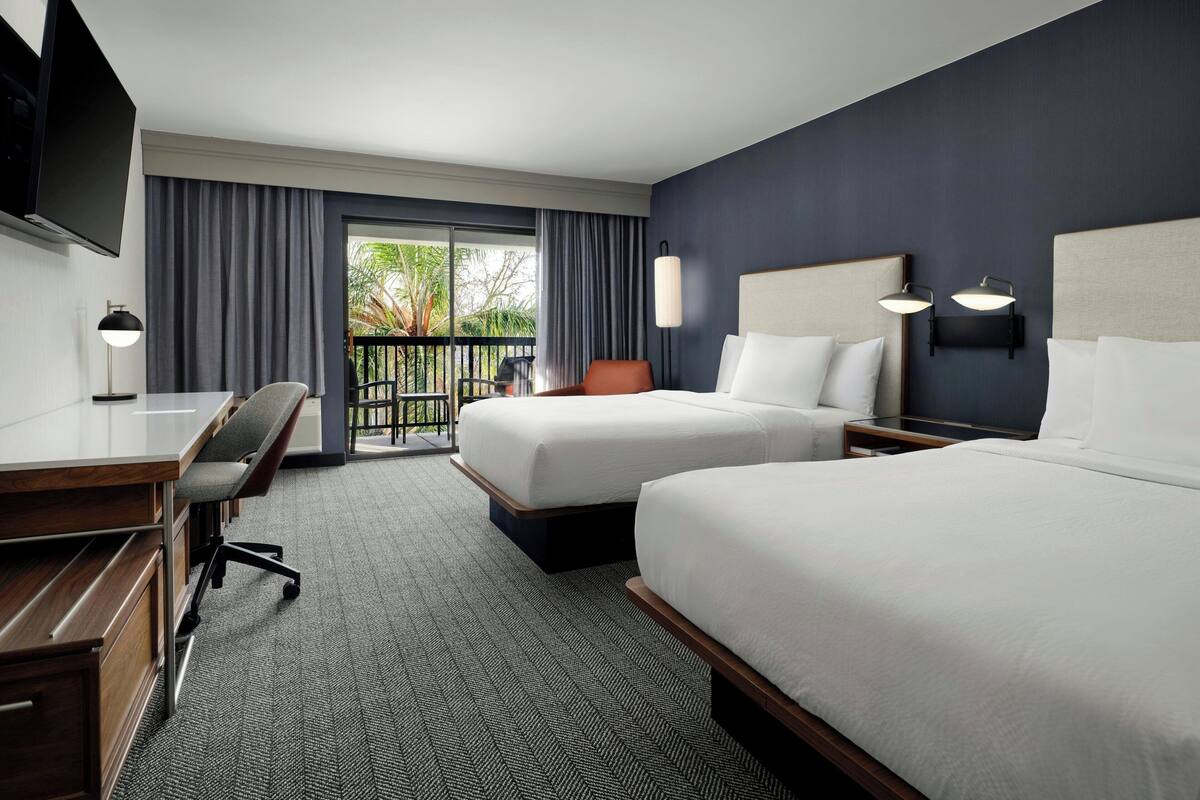 A hotel room with two beds, a desk, a chair, a TV, and a balcony with curtains.
