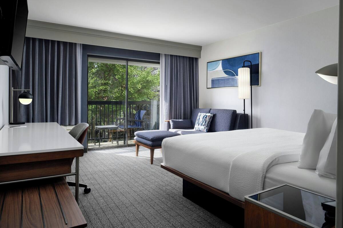 A hotel room with a bed, a desk, a chair, a small table, and a balcony with a view of trees.
