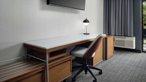 A hotel room with a wooden desk, a chair, a flat-screen TV mounted on the wall, a desk lamp, and a large window with curtains. - Courtyard by Marriott Stockton (Stockton)