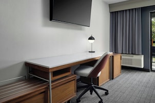 Habitación de hotel con un escritorio de madera, una silla, un televisor de pantalla plana en la pared, una lámpara de escritorio y un amplio ventanal con cortinas.