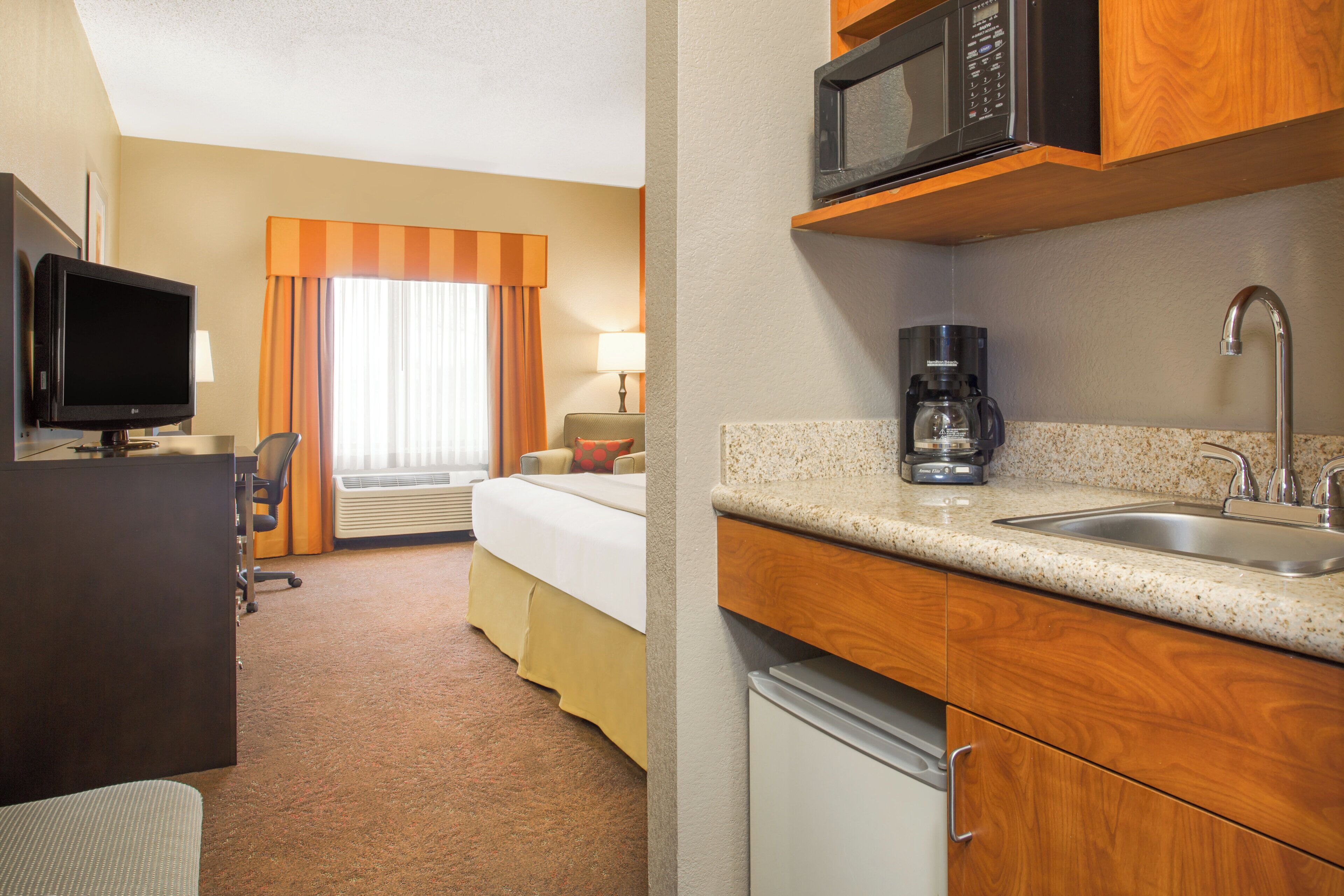 A hotel room with a bed, a desk with a TV, a kitchenette with a microwave, coffee maker, and sink.