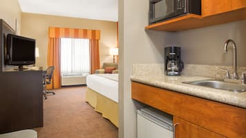 A hotel room with a bed, a desk with a TV, a kitchenette with a microwave, coffee maker, and sink.
