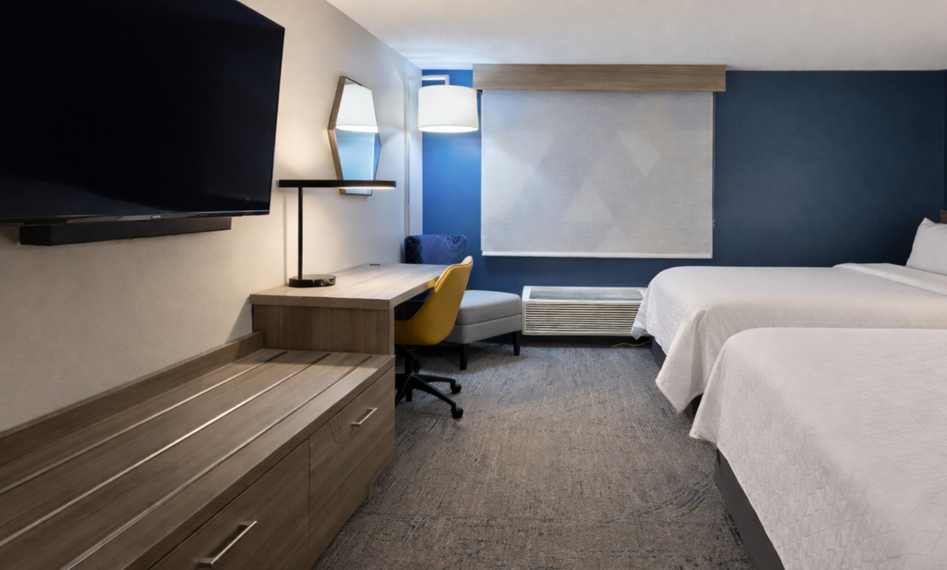 A hotel room with a bed, a desk with a chair, a flat-screen TV, a lamp, and a window with blinds.