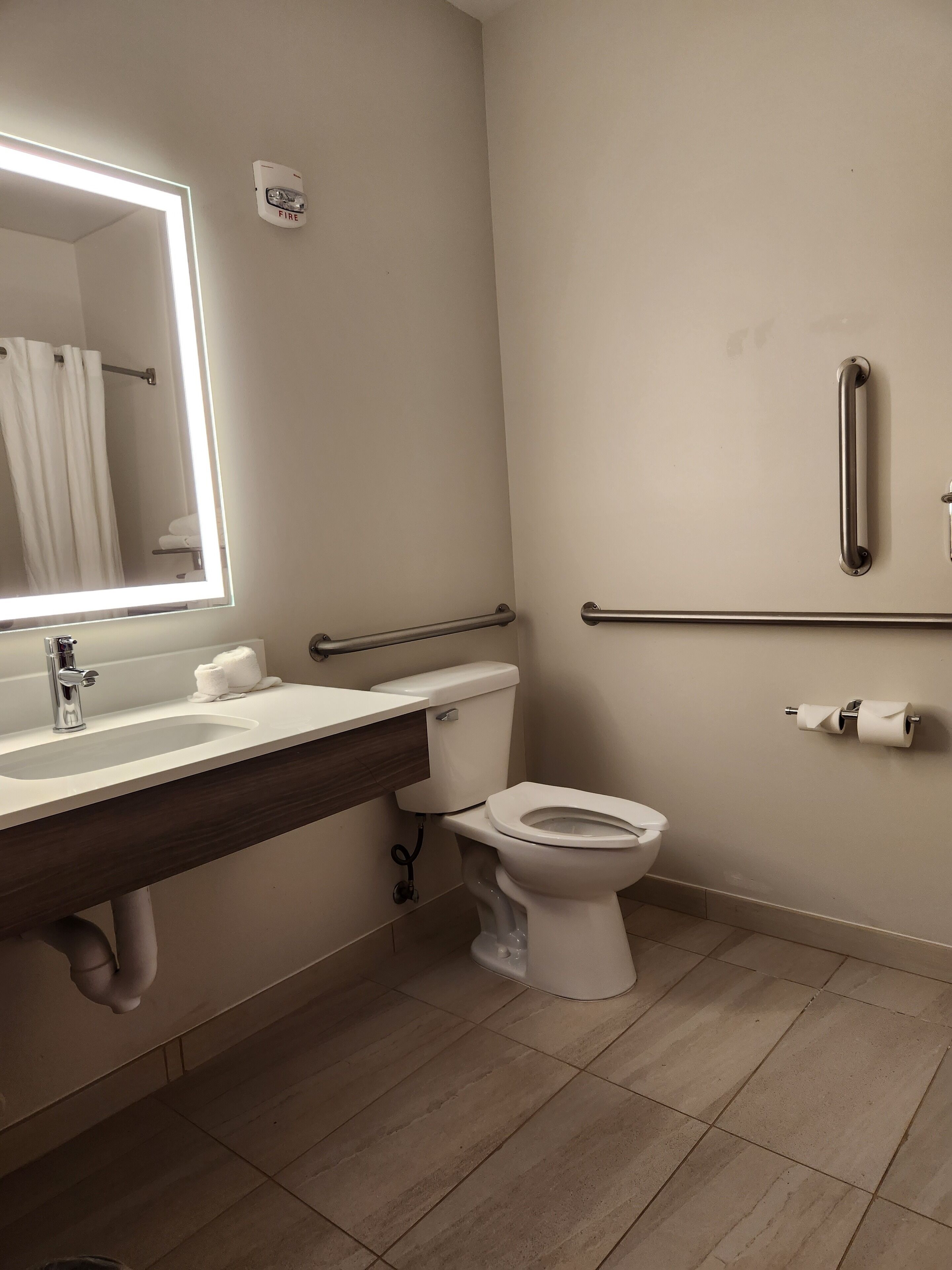 A modern bathroom with a white sink, a mirror with a light, a toilet, and a handrail.