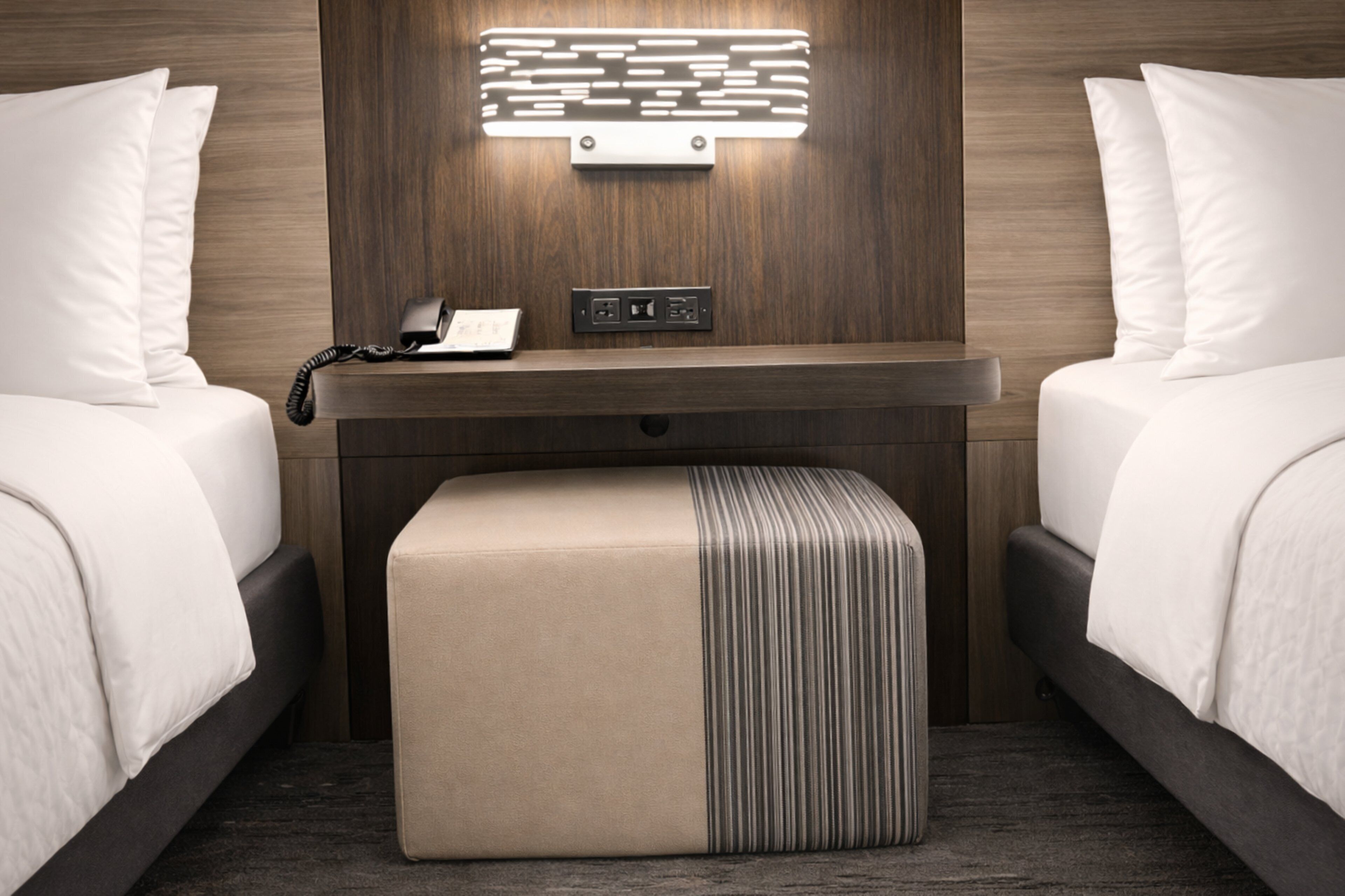 A modern hotel room with two beds, a bedside table with a phone and power outlets, and a central ottoman.