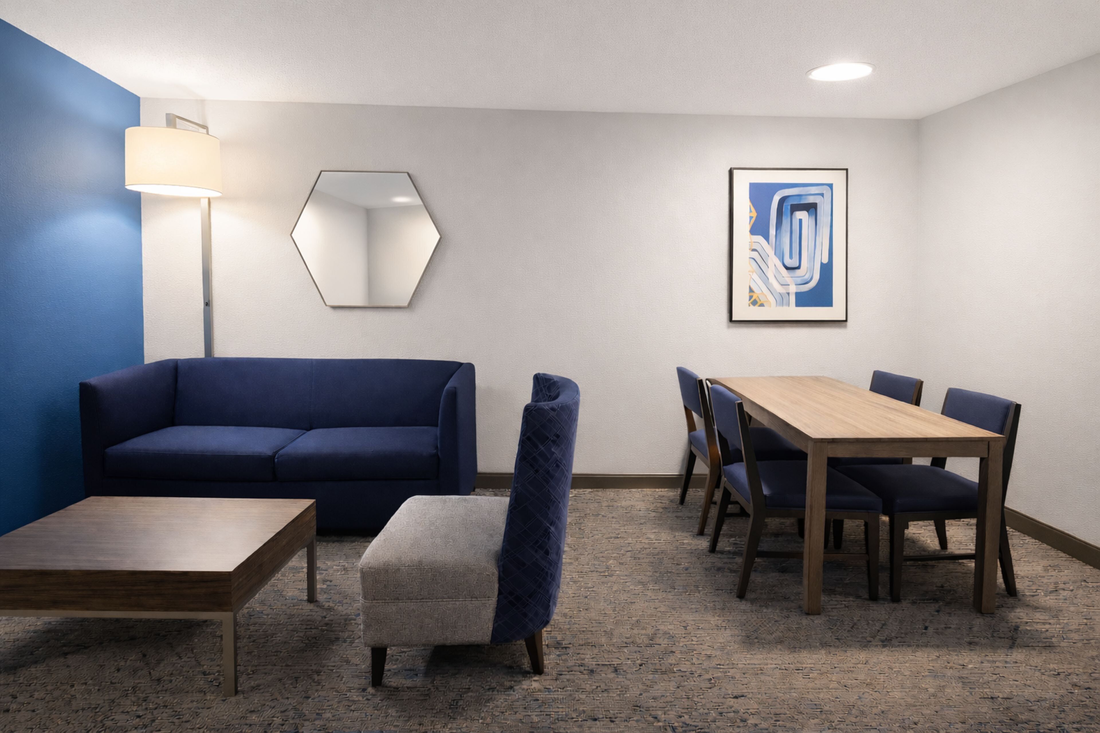Quarto moderno com sofá azul, mesa de centro de madeira, um pequeno banquinho, espelho hexagonal, mesa de jantar com cadeiras e uma pintura abstrata emoldurada.