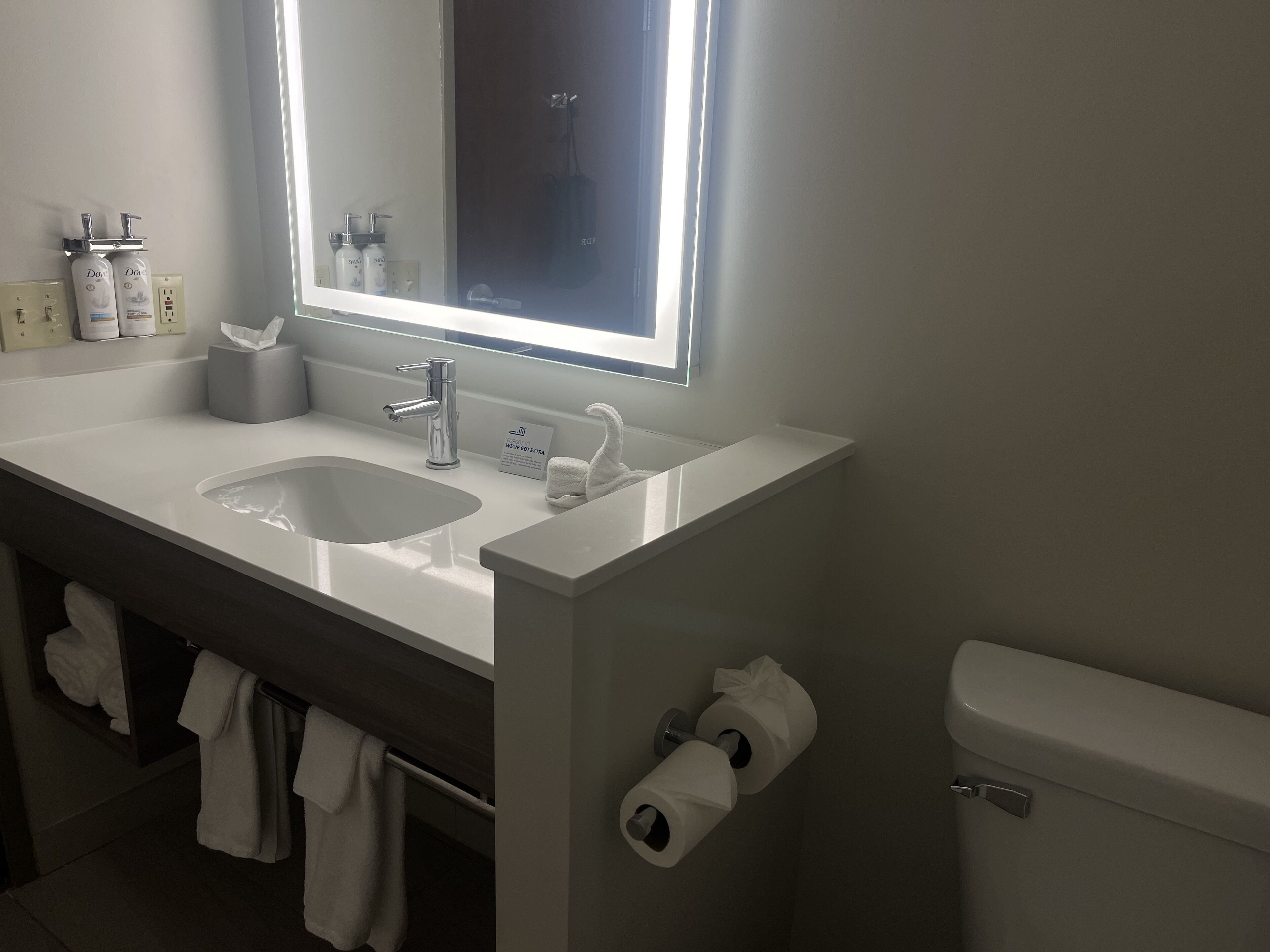 A modern bathroom with a white sink, a mirror, and a toilet. There are two rolls of toilet paper and a towel rack with towels.