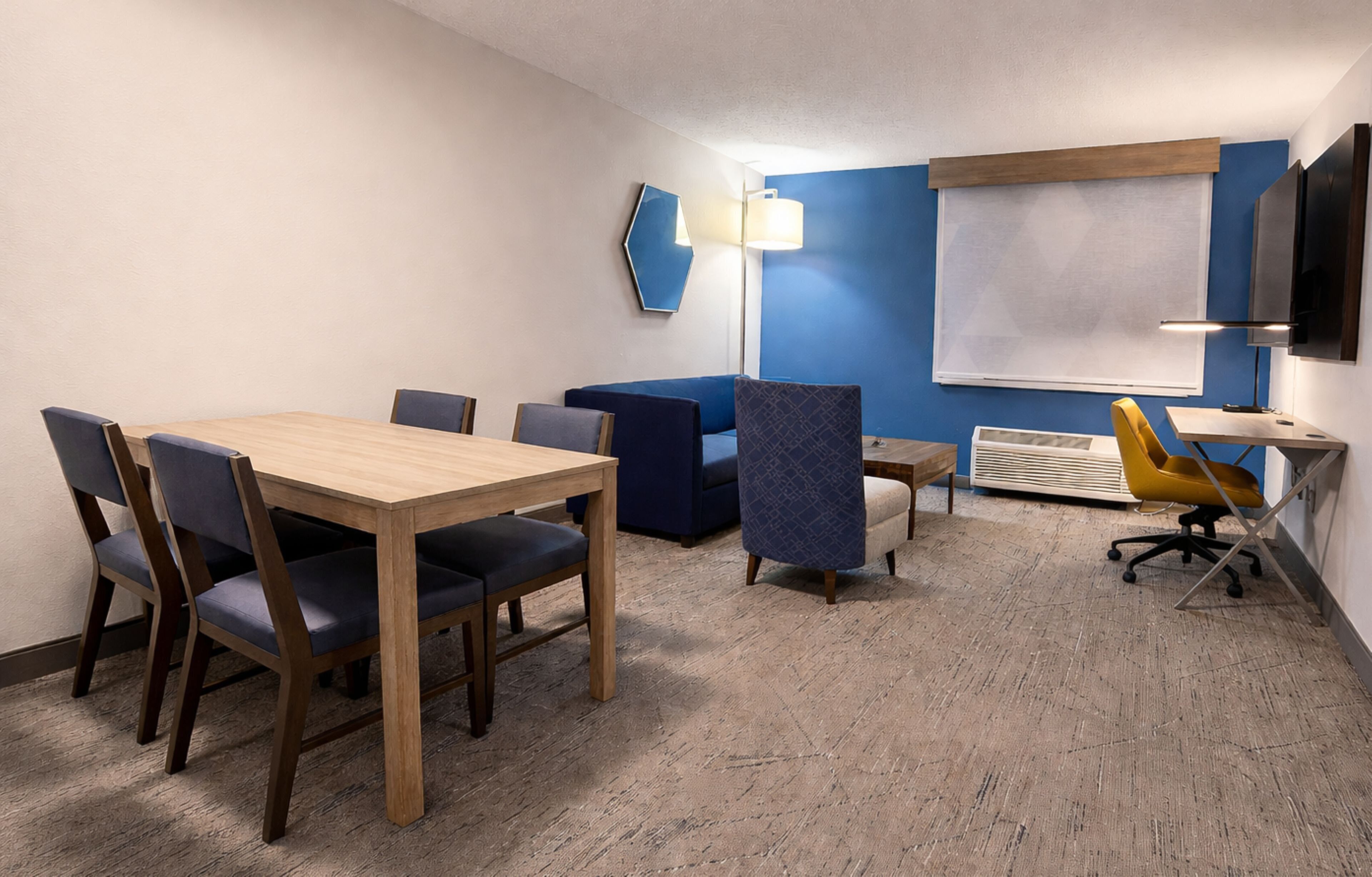 A conference room with a wooden table, blue and yellow chairs, a projector screen, and a mounted TV.