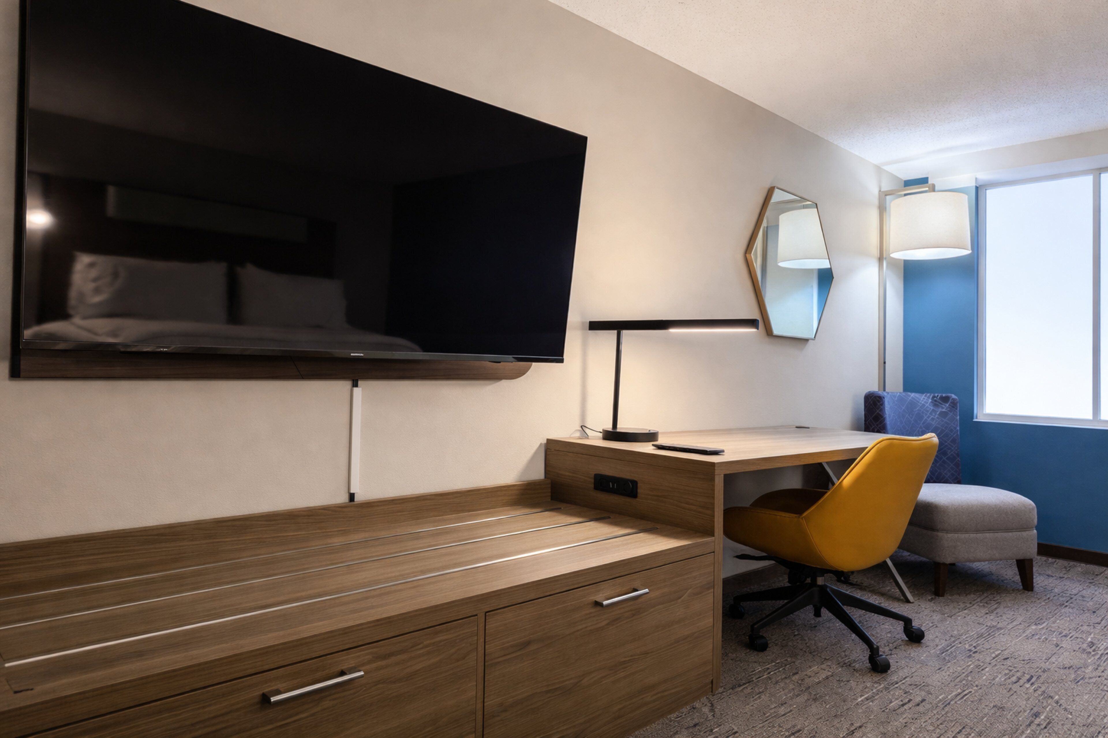 Una habitación de hotel moderna con una cama, un escritorio con una silla, un televisor de pantalla plana sobre la cama, una lámpara de noche y una ventana con vistas.