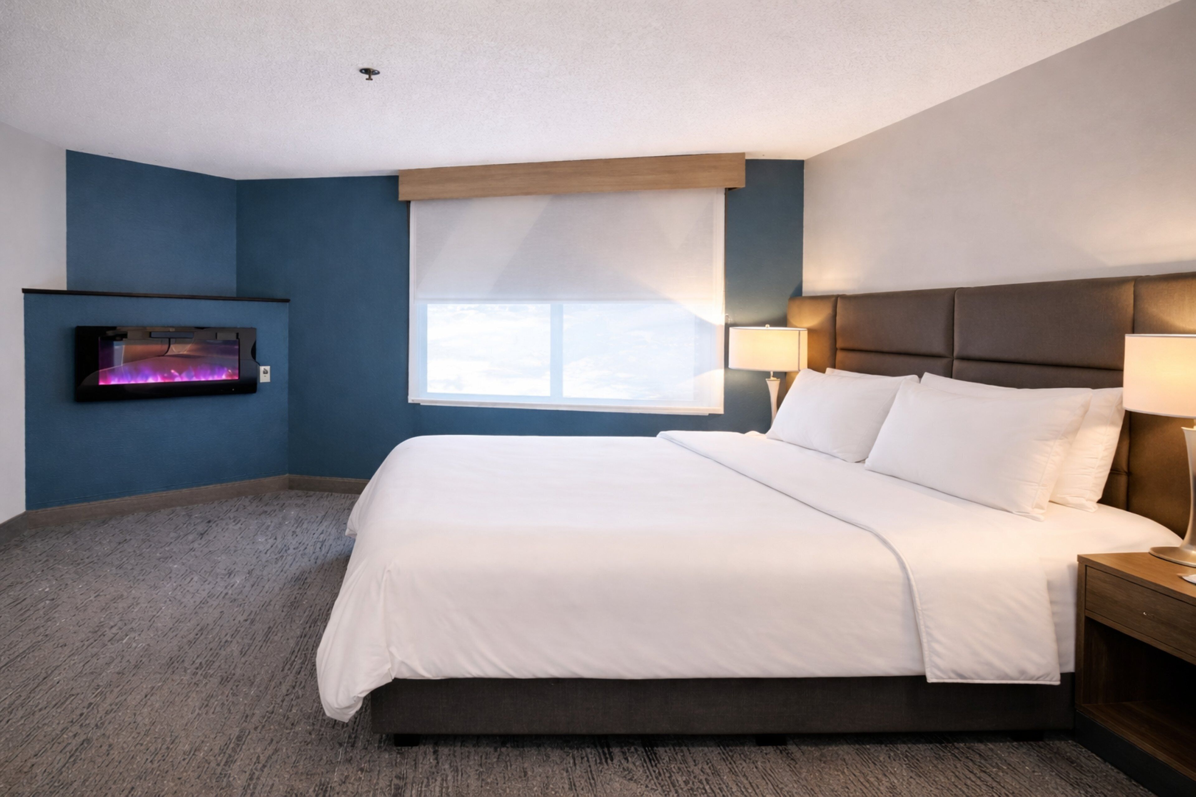 Una habitación de hotel con una cama grande, un televisor montado en la pared, una ventana con persianas y dos lámparas de noche.