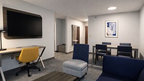 Ein Hotelzimmer mit Wandfernseher, Schreibtisch mit Stuhl, einer Couch und einem kleinen Hocker. Es gibt einen Essbereich mit Tisch und Stühlen sowie ein gerahmtes abstraktes Gemälde an der Wand.