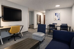 Une chambre d’hôtel équipée d’un téléviseur à écran plat fixé au mur, d’un bureau avec une chaise, d’un canapé et d’un petit pouf. On y trouve également un coin repas avec une table et des chaises, ainsi qu’un tableau abstrait encadré accroché au mur.
