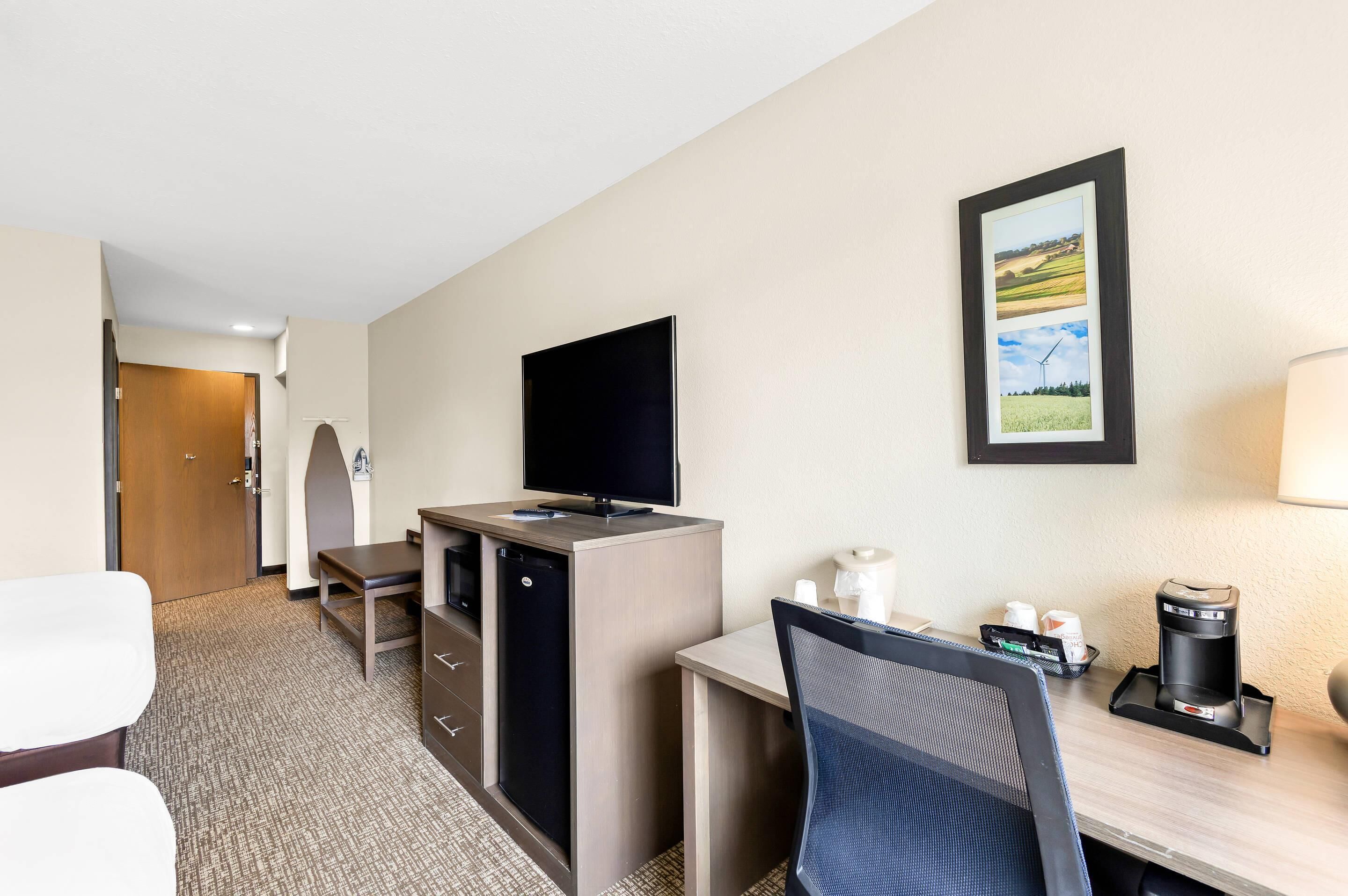 Habitación de hotel con escritorio, silla, televisor, cafetera y un cuadro de paisaje en la pared.