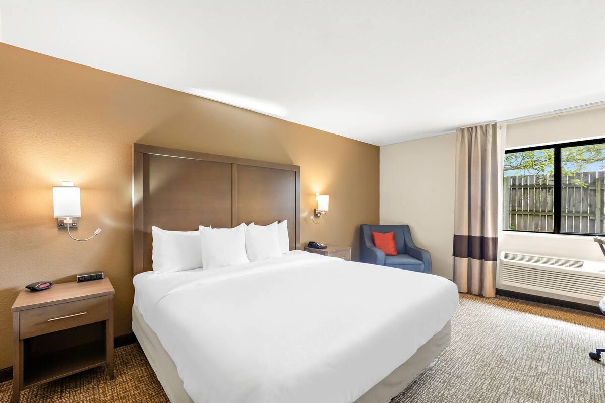 A hotel room with a large bed, bedside tables, a chair, a window with curtains, and a wall-mounted air conditioning unit.