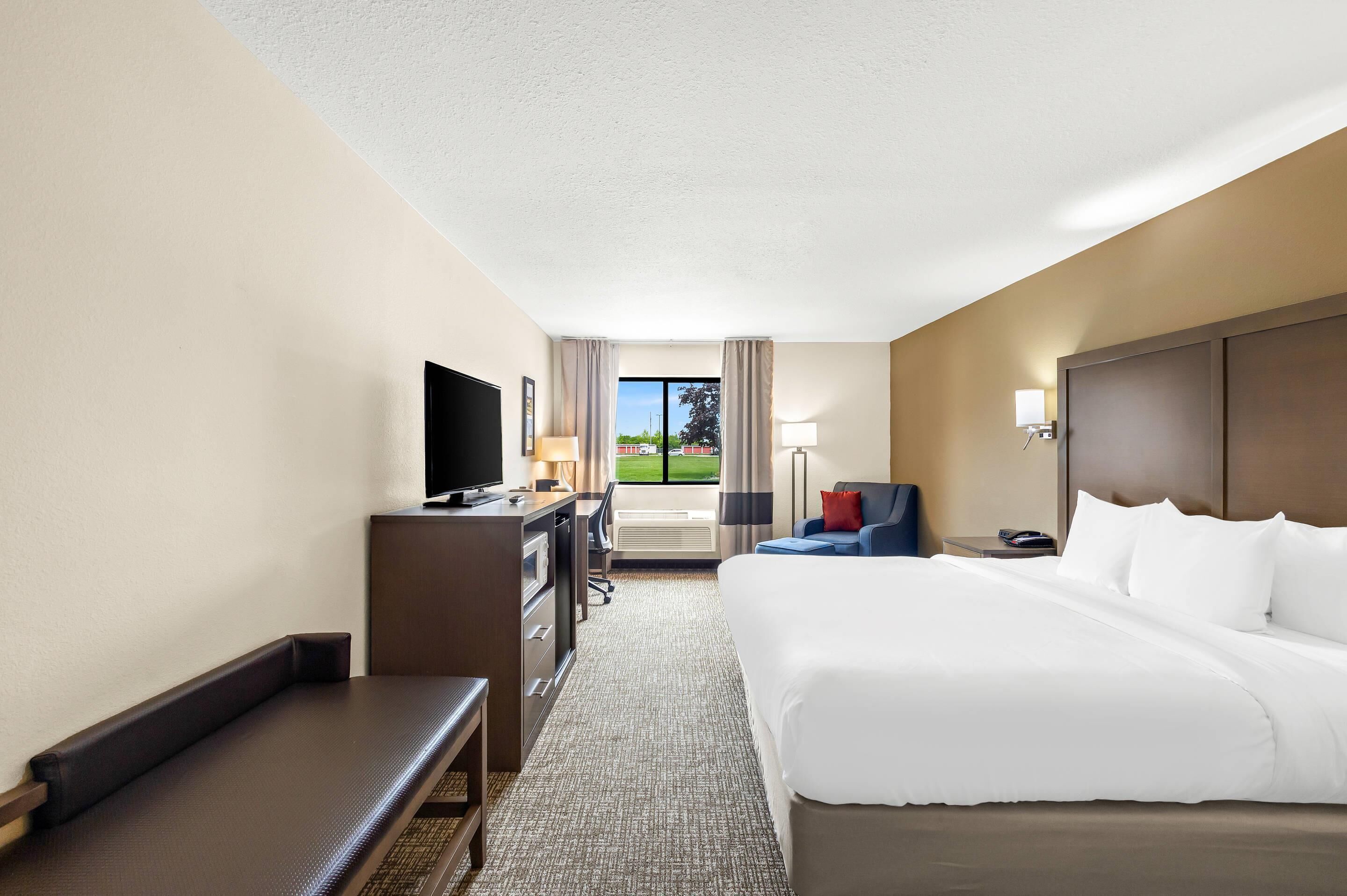 A hotel room with a large bed, a desk with a TV, a chair, a bench, and a window with a view of the outdoors.
