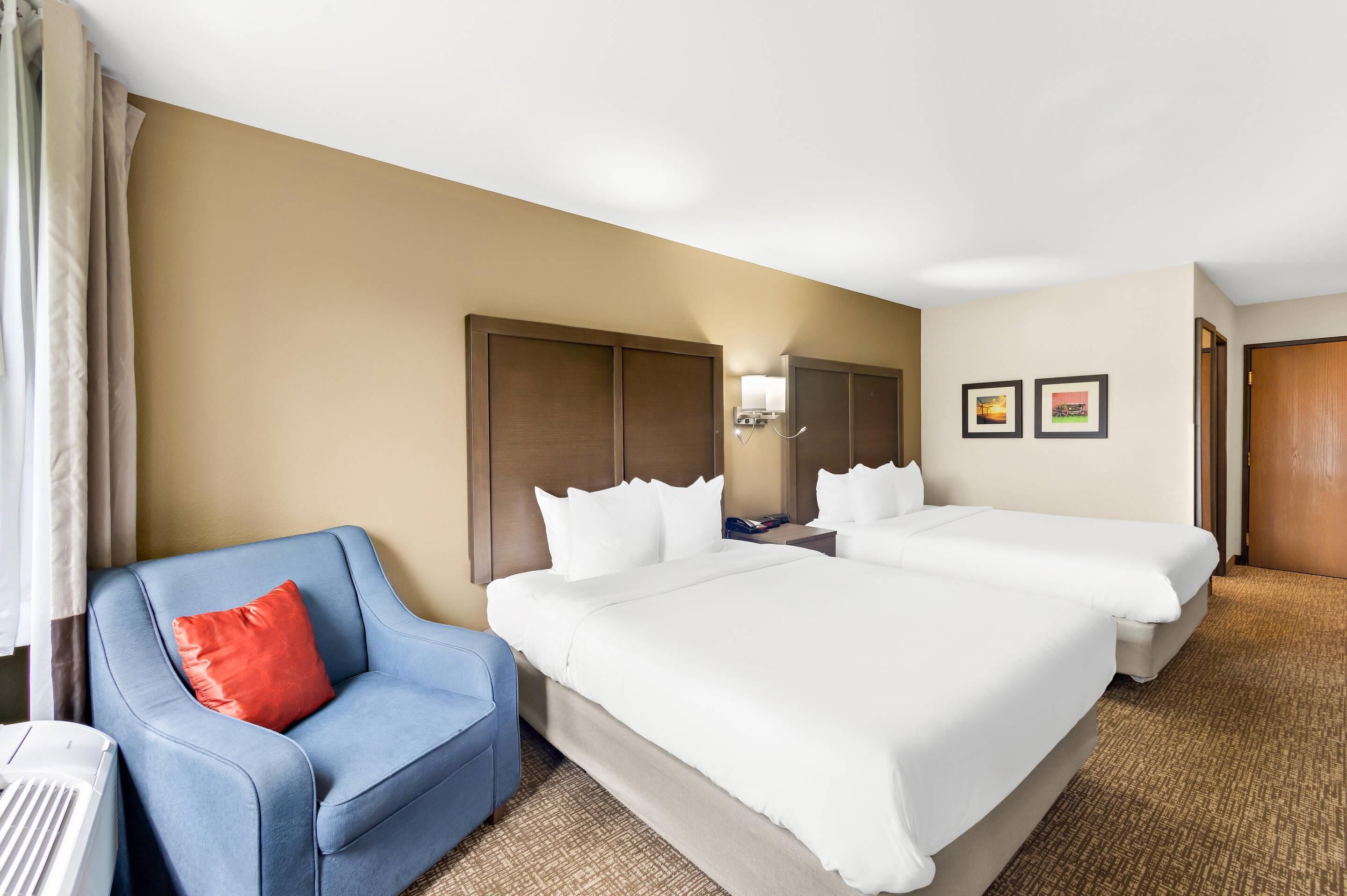 Habitación de hotel con dos camas, un sillón azul, un calentador y una lámpara fijada a la pared.