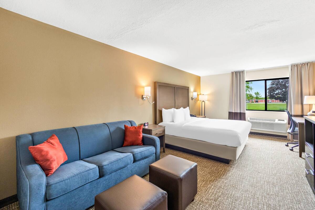 A hotel room with a bed, a blue sofa, a desk, and a view of a green field through the window.