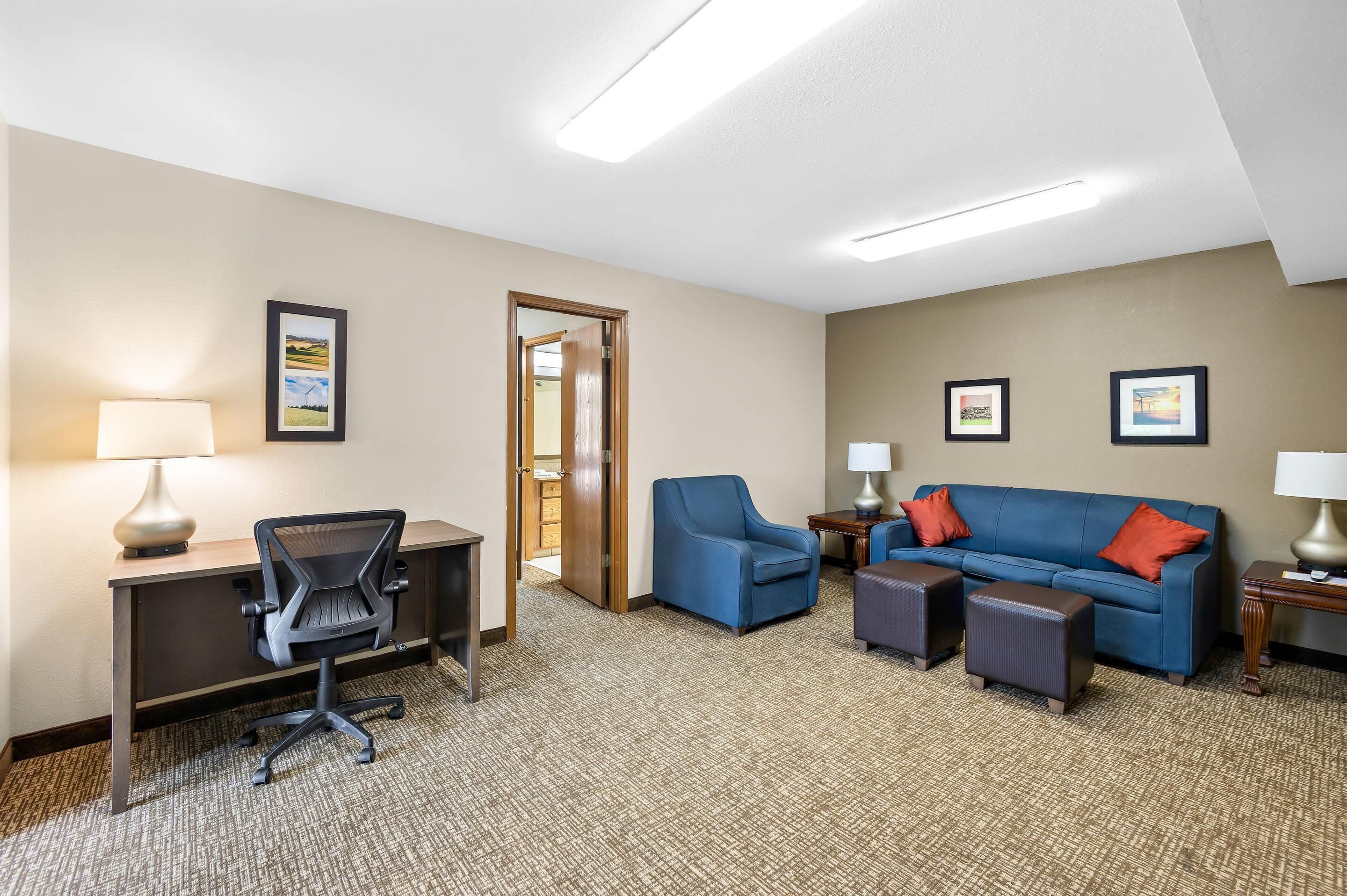 A hotel room with a desk, office chair, blue sofa, two side tables with lamps, and a carpeted floor.